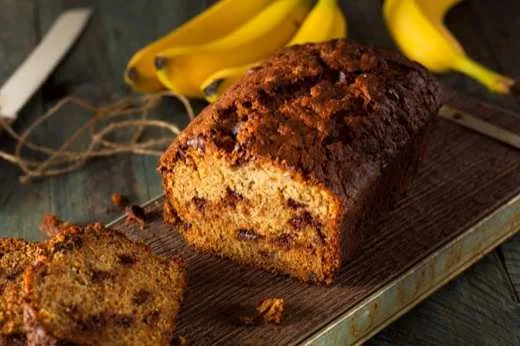 Pão de banana fatiado sobre tábua de madeira, com bananas ao fundo